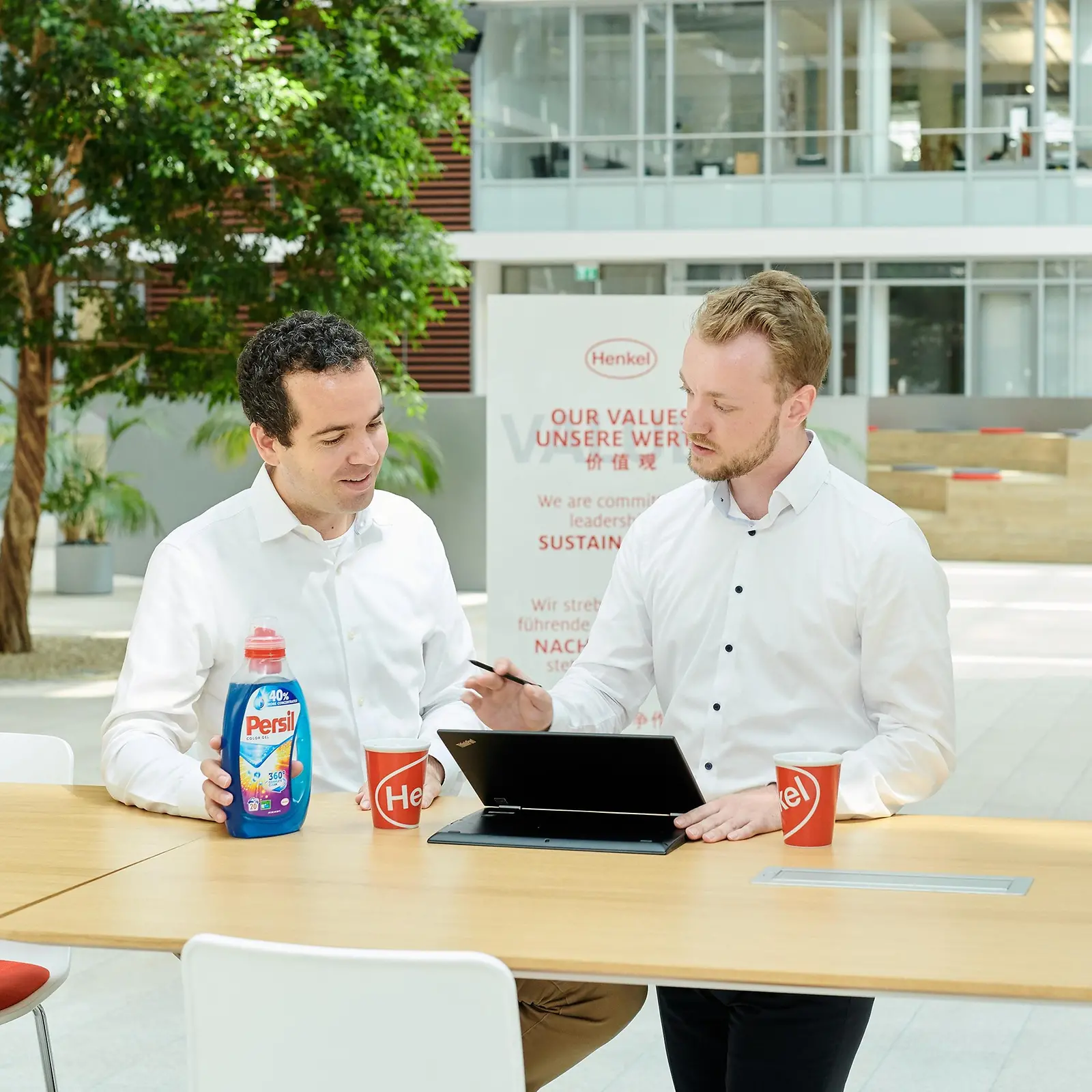 Dois homens sentados em uma mesa com um frasco de Persil e um laptop